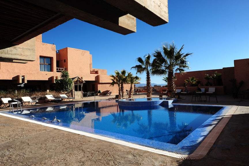 Hotel Xaluca Dades outdoor swimming pool with blue tiles surrounded by terracotta buildings and palm trees under clear blue sky