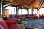 Hotel Xaluca Dades seating area with red and purple cushions on wooden benches, beige poufs, wooden tables, and a tall lamp in a rustic wooden interior