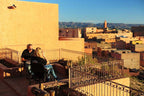 Hotel Xaluca Dades rooftop terrace with two people sitting on black chairs enjoying sunlit tan adobe walls and distant desert town