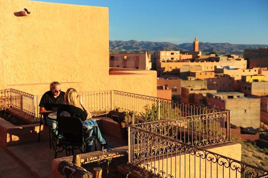 Hotel Xaluca Dades rooftop terrace with two people sitting on black chairs enjoying sunlit tan adobe walls and distant desert town