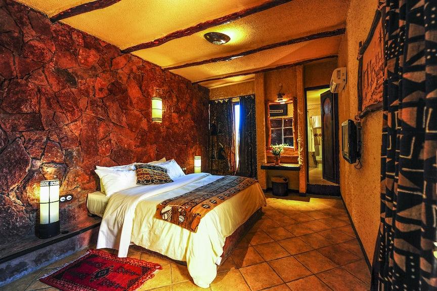 Hotel Xaluca Dades room with stone textured wall, warm yellow lighting, tiled floor, white bed with patterned throw and cushions