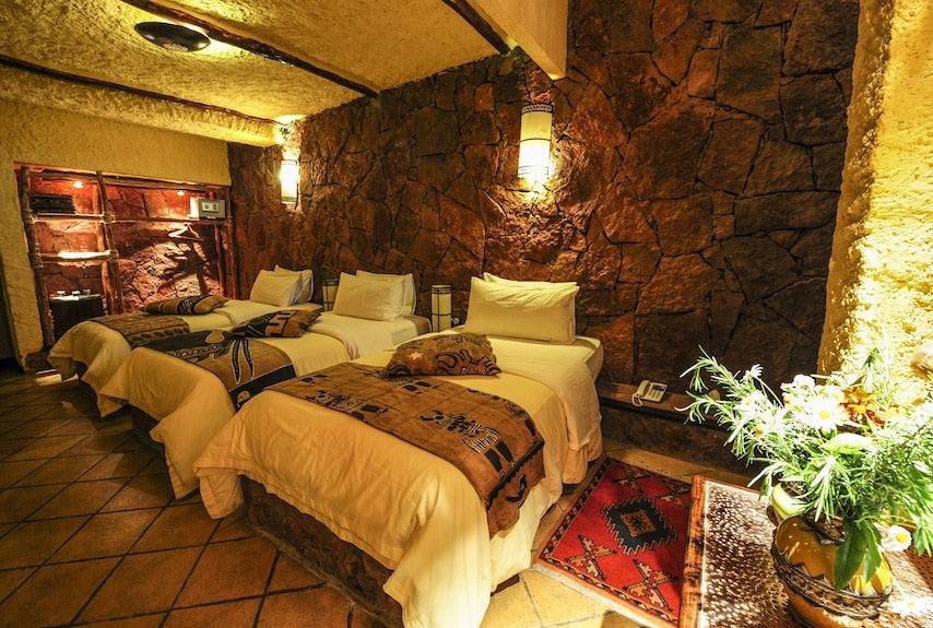 Hotel Xaluca Dades room with three single beds featuring white linens, brown patterned throws, stone walls, and terracotta tiled floor