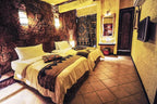 Hotel Xaluca Dades twin bedroom with white bedding, decorative brown and black patterned runners, stone wall, and traditional Moroccan decor
