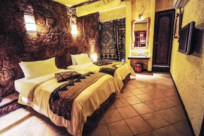 Hotel Xaluca Dades twin bedroom with white bedding, decorative brown and black patterned runners, stone wall, and traditional Moroccan decor