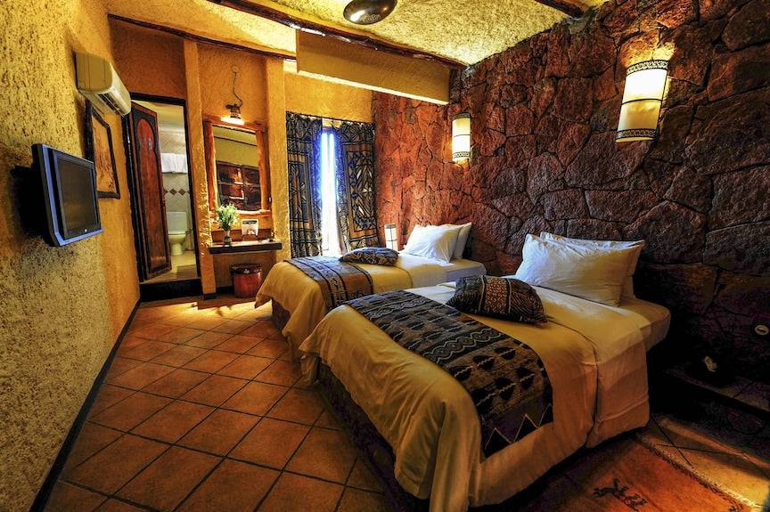 Hotel Xaluca Dades twin beds with white linens, patterned throws, stone accent wall, warm Moroccan-style lighting