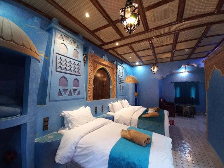 Hotel Yasmina Merzouga room with blue Moroccan-style walls, wooden ceiling, twin beds with white linens and teal runners, and intricate decorative woodwork