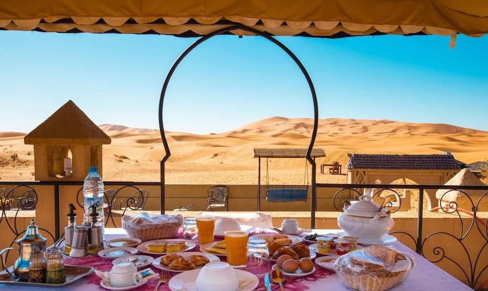 Hotel Yasmina Merzouga outdoor dining table with breakfast spread, orange juice, bread, eggs, and desert dunes view