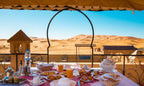 Hotel Yasmina Merzouga outdoor dining table with breakfast spread, orange juice, bread, eggs, and desert dunes view
