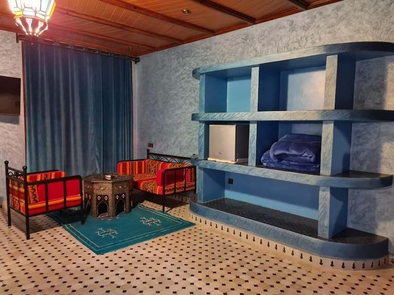 Hotel Yasmina Merzouga cozy sitting area with blue walls, patterned red cushions, a small wooden table, and blue carpet