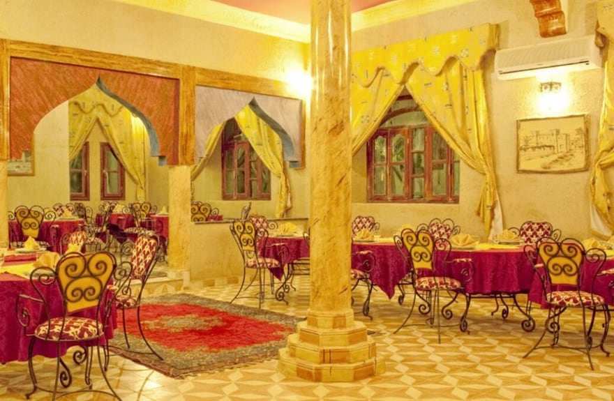Hotel Yasmina Merzouga dining area with yellow walls, wrought iron chairs with red patterned cushions, and red tablecloths