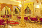 Hotel Yasmina Merzouga dining area with yellow walls, wrought iron chairs with red patterned cushions, and red tablecloths