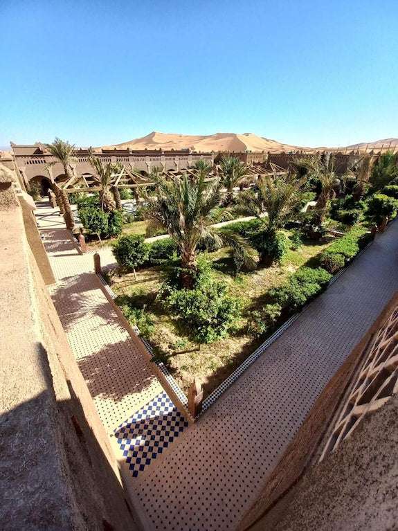 Hotel Yasmina Merzouga showcasing a lush green garden courtyard with palm trees and desert dunes in the background under clear blue sky