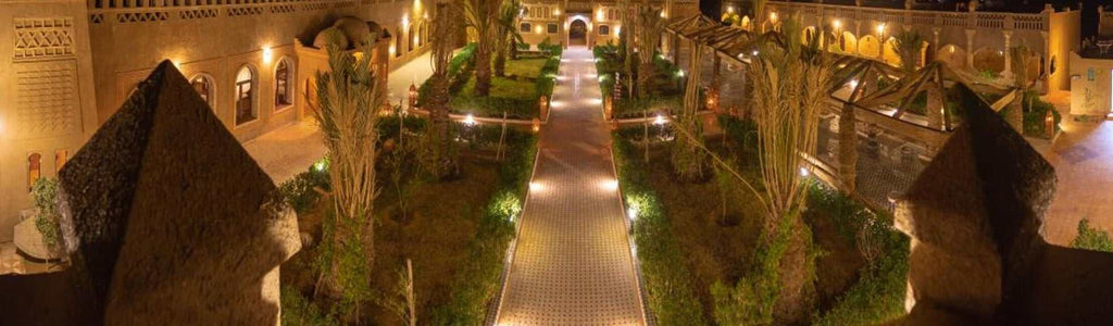 Hotel Yasmina Merzouga courtyard at night featuring illuminated tiled walkway, palm trees, and traditional Moroccan architecture