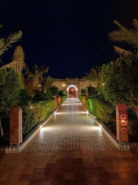 Hotel Yasmina Merzouga illuminated entrance pathway at night with lush green plants and ornate lanterns lining the tiled walkway