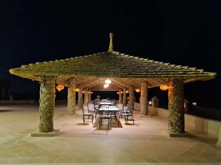 Hotel Yasmina Merzouga outdoor seating area at night featuring a large woven palm-thatched roof supported by column pillars with hanging lanterns