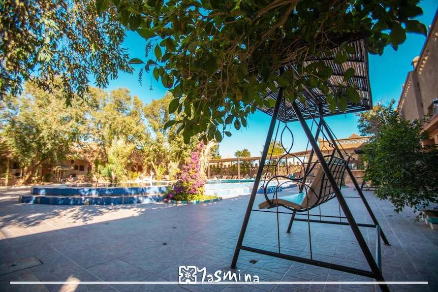 Outdoor area at Hotel Yasmina Merzouga featuring a black metal swing chair under leafy trees beside a tiled pool and lush greenery