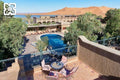 Hotel Yasmina Merzouga outdoor pool area with terracotta tiles, wrought iron balcony, pink cushioned chairs, and desert dunes backdrop.