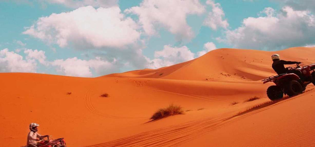 Hotel Yasmina Merzouga adventure showing two people riding red all-terrain vehicles on vast orange sand dunes under a partly cloudy blue sky
