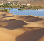Hotel Yasmina Merzouga seen beyond golden sand dunes with a blue oasis and green vegetation in desert landscape