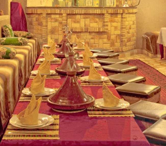 Hotel Yasmina Merzouga traditional low dining setup with brown tagine pots, folded yellow napkins, and cushioned seats on a red carpet