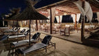 Kal Tenere Agafay luxury desert camp outdoor lounge area at night with wooden furniture, fabric chairs, and thatched umbrellas