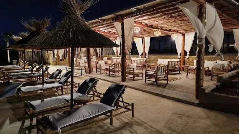 Kal Tenere Agafay luxury desert camp outdoor lounge area at night with wooden furniture, fabric chairs, and thatched umbrellas