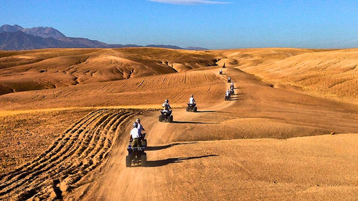 Kal Tenere Agafay desert scene with riders on quad bikes traveling along a dirt path through golden sandy dunes under a clear blue sky