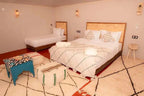 Kal Tenere Agafay luxury desert camp bedroom with double and single beds, beige walls, patterned cushions and rugs, and a blue side table