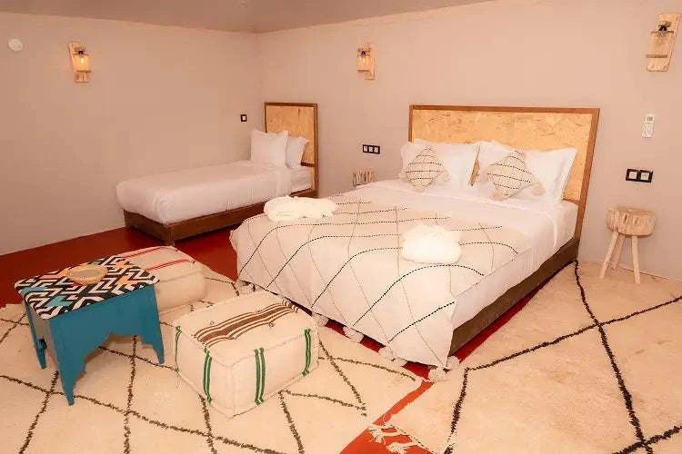 Kal Tenere Agafay luxury desert camp bedroom with double and single beds, beige walls, patterned cushions and rugs, and a blue side table