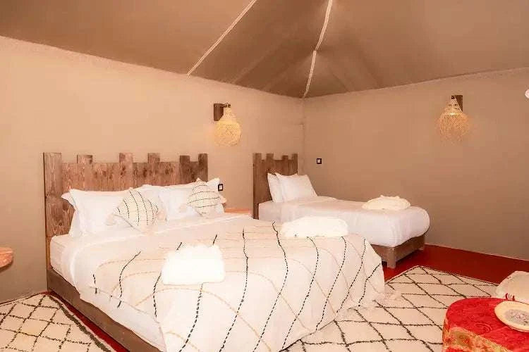 Kal Tenere Agafay luxury desert camp bedroom with beige walls, wooden headboards, white bedding with black pattern, and decorative rugs