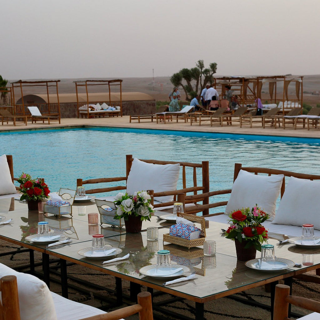 Kal Tenere Agafay luxury desert camp outdoor dining table with white cushions, floral centerpieces beside a turquoise swimming pool