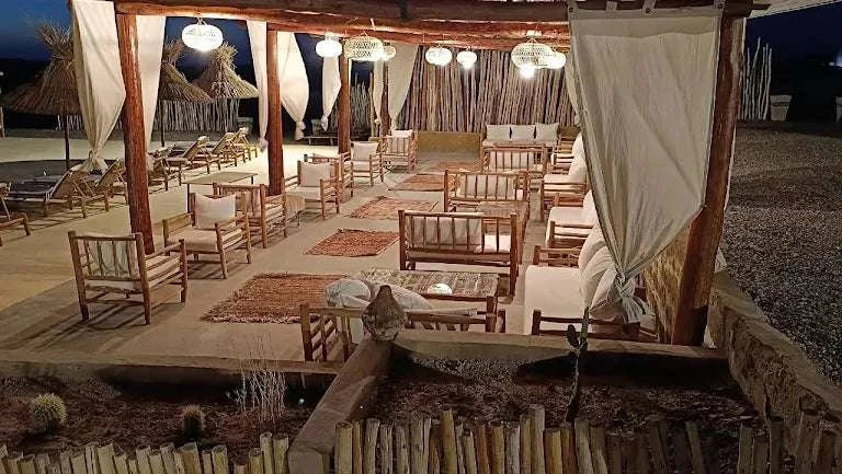 Kal Tenere Agafay outdoor luxury desert camp seating area with wooden chairs, beige cushions, rugs, and hanging lanterns
