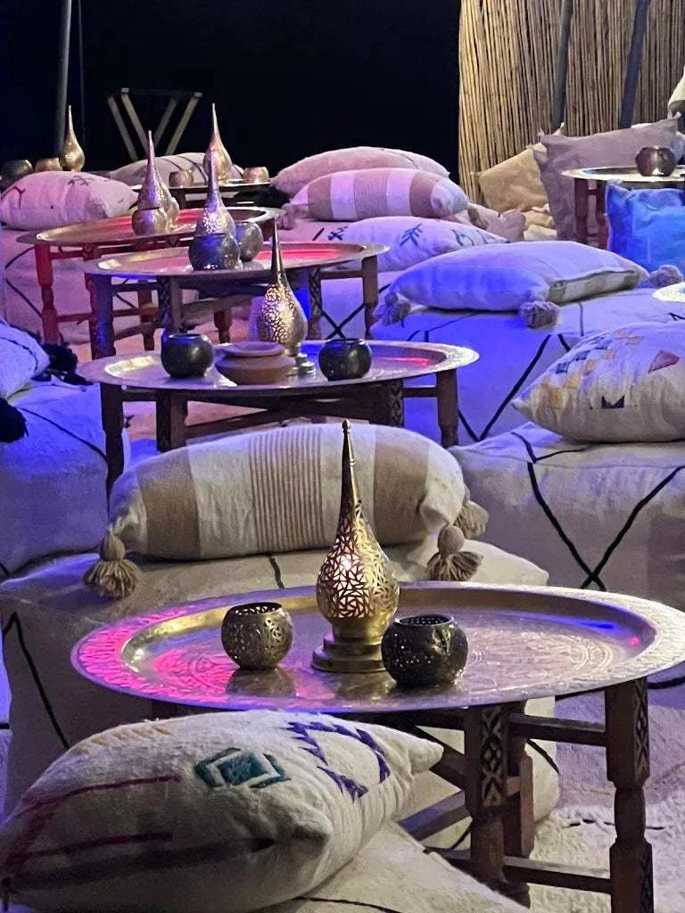 Kal Tenere Agafay luxury desert camp low wooden tables with brass lanterns and cushions with Moroccan patterns under ambient lighting