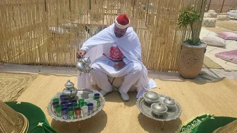 Kal Tenere Agafay traditional Moroccan tea ceremony setup with a man in white and red attire pouring tea on a woven mat