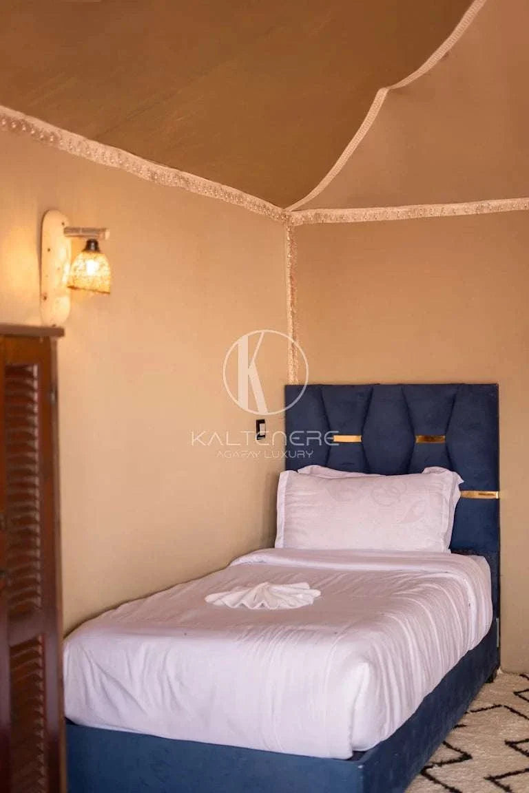 Kal Tenere Agafay single bed with white linens and navy upholstered headboard inside beige tent room with warm lamp