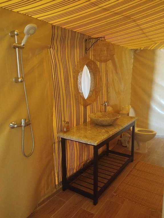 Kalimba Luxury Desert Camp bathroom with beige canvas walls, striped fabric accents, stone sink on granite countertop, wicker mirror, and shower