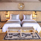 Kalimba Luxury Desert Camp bedroom with two single beds featuring white linens, grey cushions, woven wood headboard, and rustic bench