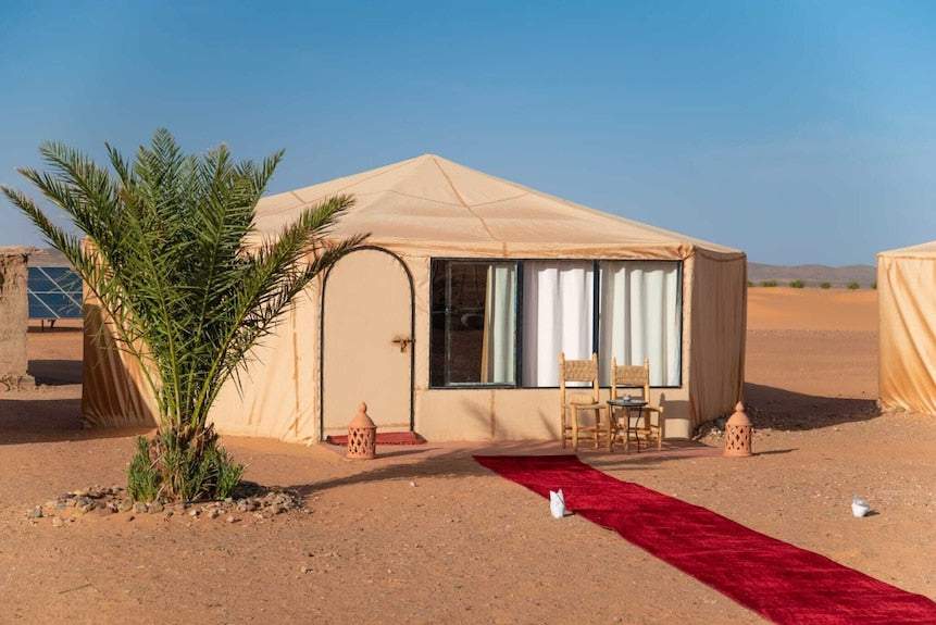 Kalimba Luxury Desert Camp beige fabric tent with arched door, window with white curtains, two chairs and red carpet on desert sand