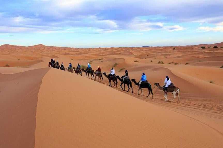 Kalimba Luxury Desert Camp camel caravan with riders crossing golden sand dunes under a cloudy blue sky