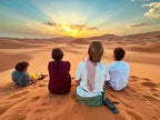 Four people sitting on sand dunes watching a vibrant desert sunset over golden Sahara dunes at Kalimba Luxury Desert Camp