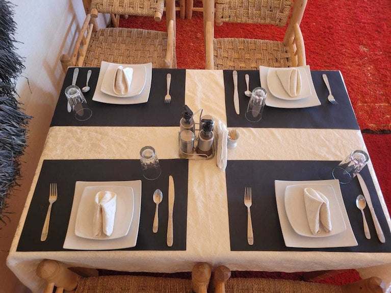Kalimba Luxury Desert Camp dining table set with black and beige tablecloth, square white plates, glass cups, and silver cutlery on red carpet