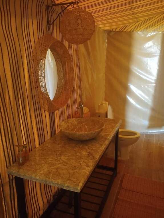 Kalimba Luxury Desert Camp bathroom featuring striped fabric walls, stone sink, round wicker mirror, and white toilet