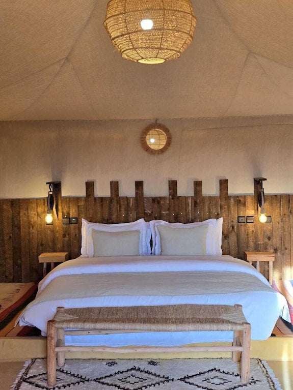 Kalimba Luxury Desert Camp interior with white bedding, wooden headboard, bedside lamps, and woven pendant light above the bed