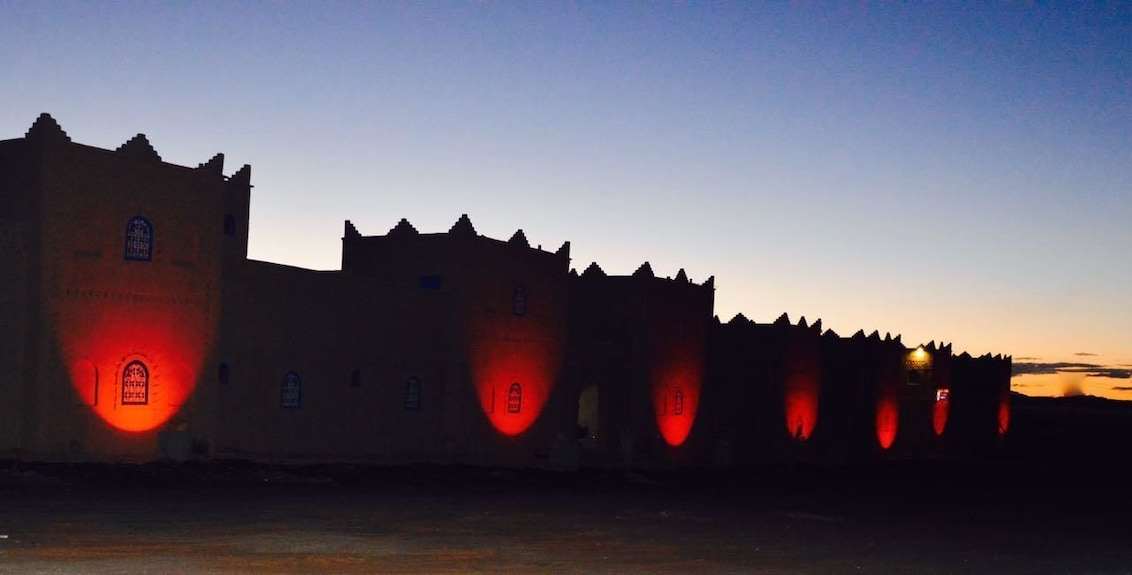 Kasbah Hotel Bar in Merzouga Morocco silhouette at dusk with traditional red mudbrick architecture and lit windows under a twilight sky
