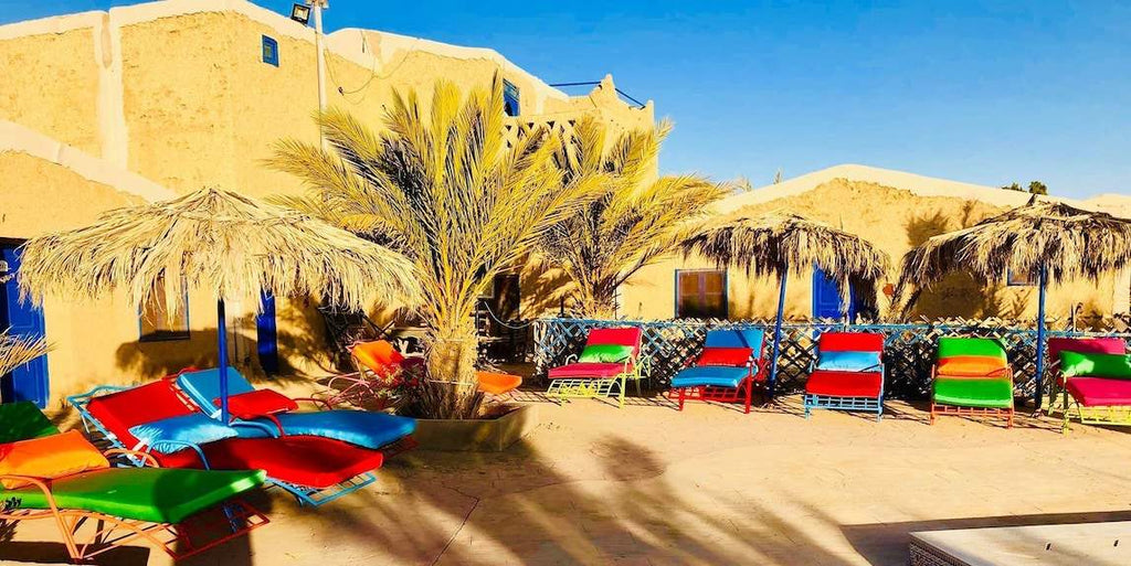 Kasbah Hotel Bar outdoor seating with colorful cushioned lounge chairs and thatched umbrellas against sandy adobe walls in Merzouga