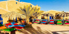 Kasbah Hotel Bar outdoor seating with colorful cushioned lounge chairs and thatched umbrellas against sandy adobe walls in Merzouga