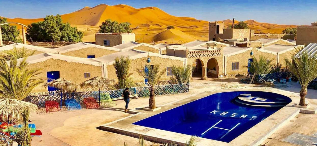 Kasbah Hotel Bar outdoor pool area with blue water, sandy beige walls, palm trees, and golden desert dunes in background
