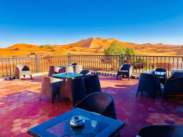 Kasbah Hotel Bar terrace with black wicker chairs and tables overlooking golden Sahara Desert sand dunes under blue sky