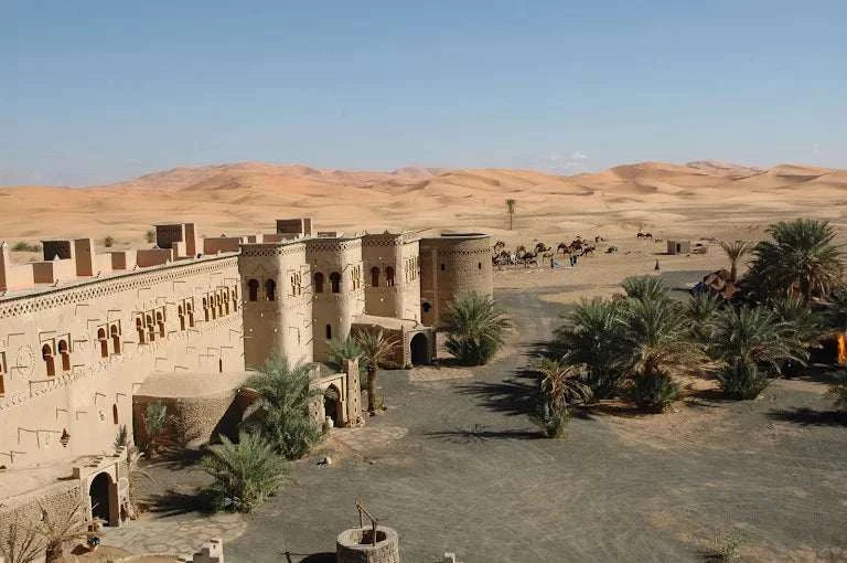 Kasbah Hotel Tombouctou beige desert kasbah-style building with decorative windows surrounded by palm trees and sand dunes