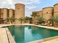 Kasbah Hotel Tombouctou exterior showing rustic beige stone walls and round towers surrounding a clear turquoise swimming pool with lounge chairs and palm trees
